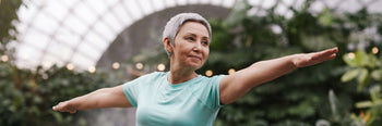 Elderly woman performing a yoga warrior pose in a lush green indoor botanical garden